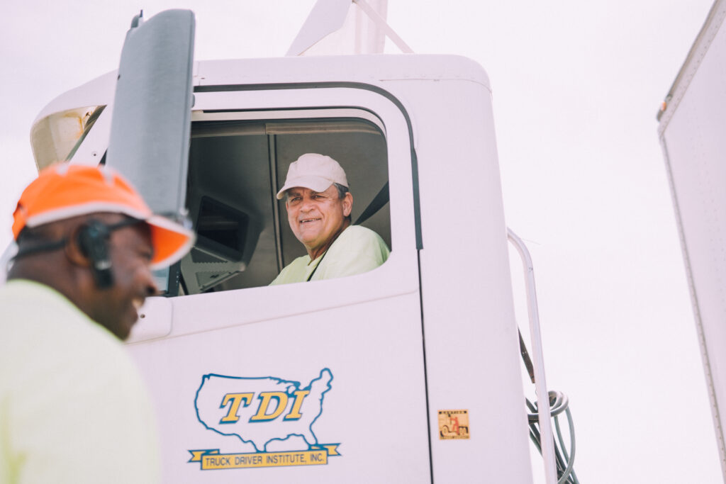 A TDI instructor sits in the cab of a parked semi truck with the TDI logo on the door, looking down. Another instructor in the foreground laughs.